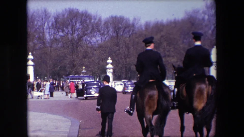 1967 London frame showing mounted uniformed riders from behind, a uniformed person walking ahead, pedestrians near parked or moving cars, and bare trees beyond the roadway.