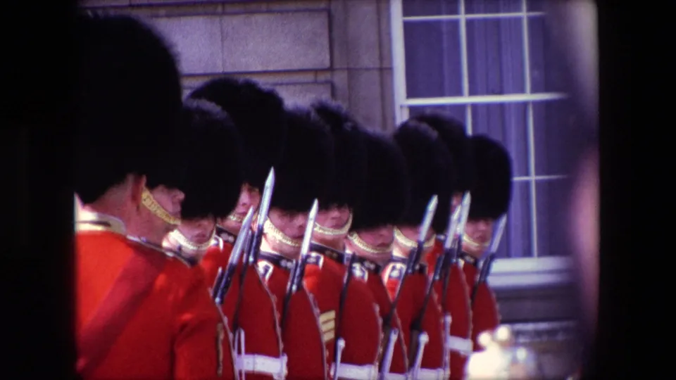 London 1967 frame showing a line of ceremonial guards in red uniforms, dark hats, and white belts, with rifles and bayonets set against a stone building facade.