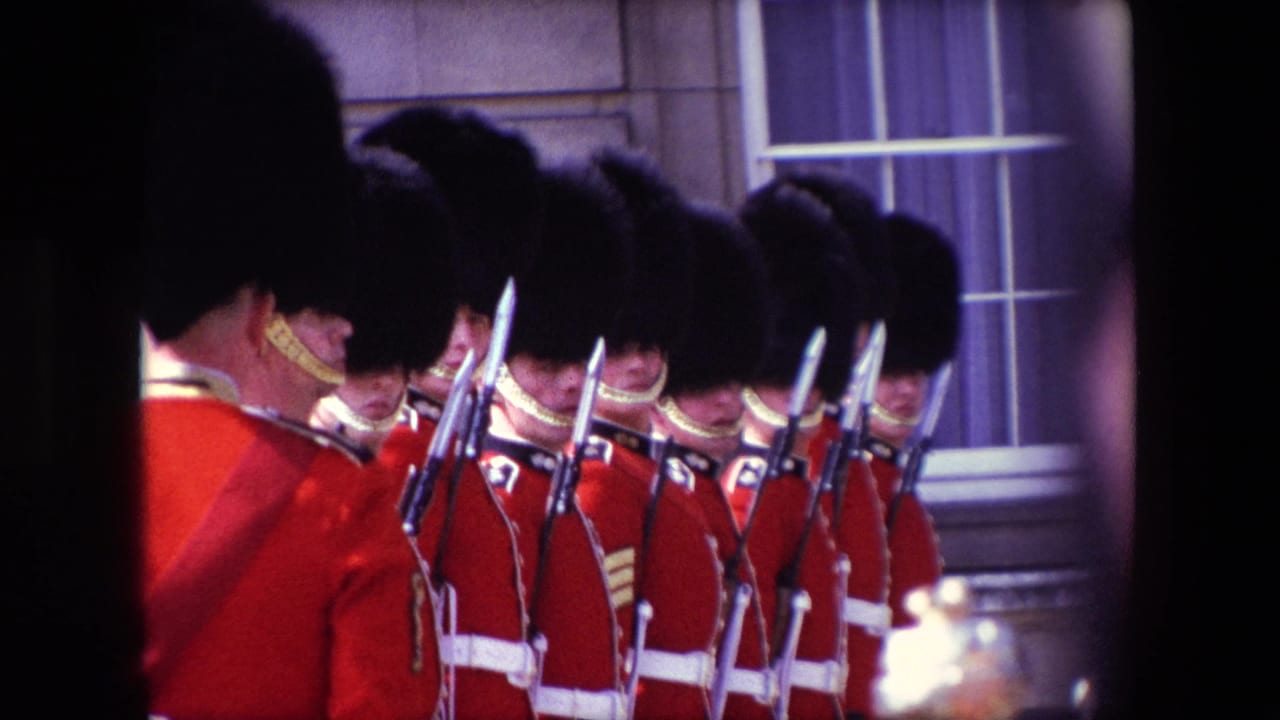 1967 London home-movie frame showing ceremonial guards in red tunics and tall black caps standing in formation with bayonets near a stone facade.
