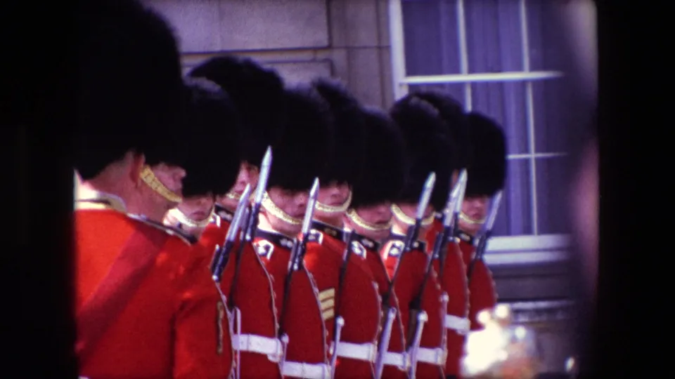 1967 London home-movie frame showing ceremonial guards in red tunics and tall black caps standing in formation with bayonets near a stone facade.