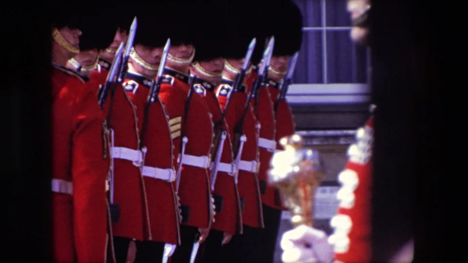 Close 1967 London home-movie view of red-coated ceremonial guards standing shoulder to shoulder, with bayonets and building windows behind them.