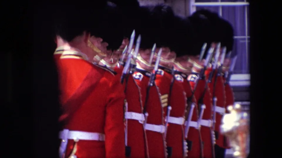A 1967 London home-movie frame shows a line of ceremonial guards in red uniforms and tall black caps standing with bayonets, partly blocked by a guard in the foreground.