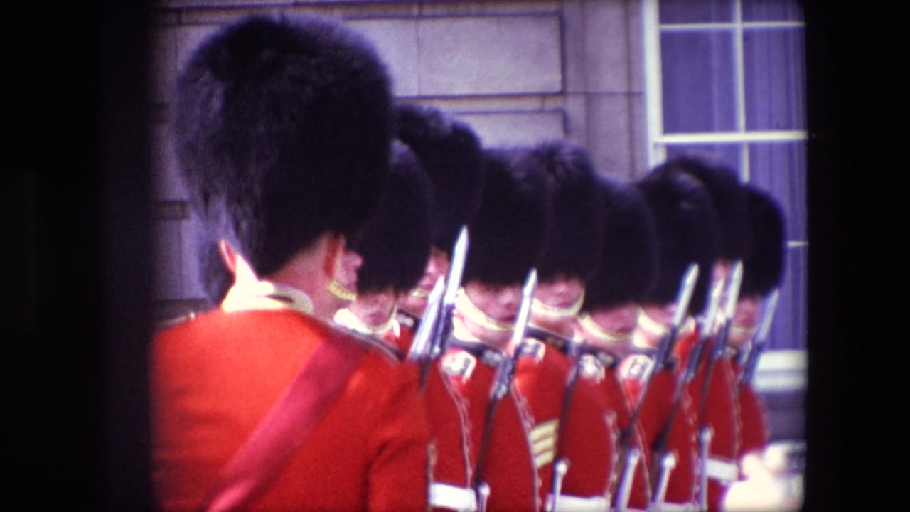 Uniformed British guards in red coats and black caps stand with rifles near a stone building.