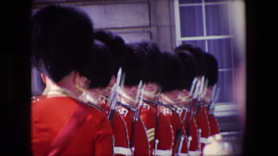 1967 London home-movie frame showing a clustered guard formation with red tunics, tall black caps, rifles, and a building window behind them.
