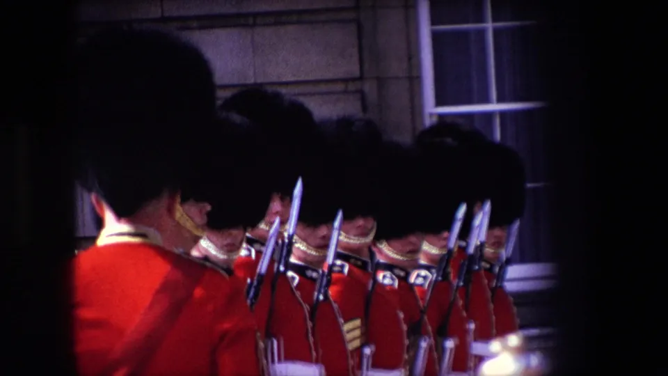 Darker 1967 London frame showing ceremonial guards in red coats and tall black caps holding rifles with bayonets near a stone exterior wall.
