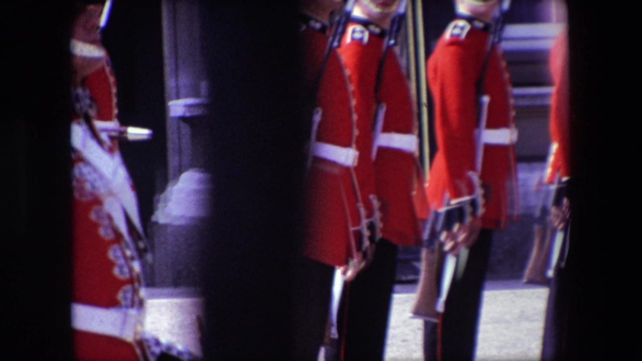 Line of ceremonial guards in red uniforms holding rifles near a stone building.