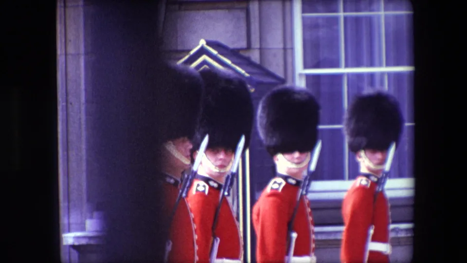 1967 London home-movie frame with three ceremonial guards in red tunics, tall dark hats, and rifles standing before a stone facade and window.