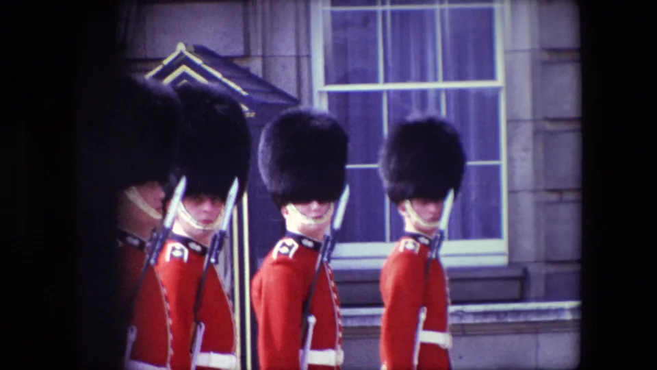 1967 London frame showing three ceremonial guards in red tunics and bearskin-style hats standing in a row with rifles outside a stone building.