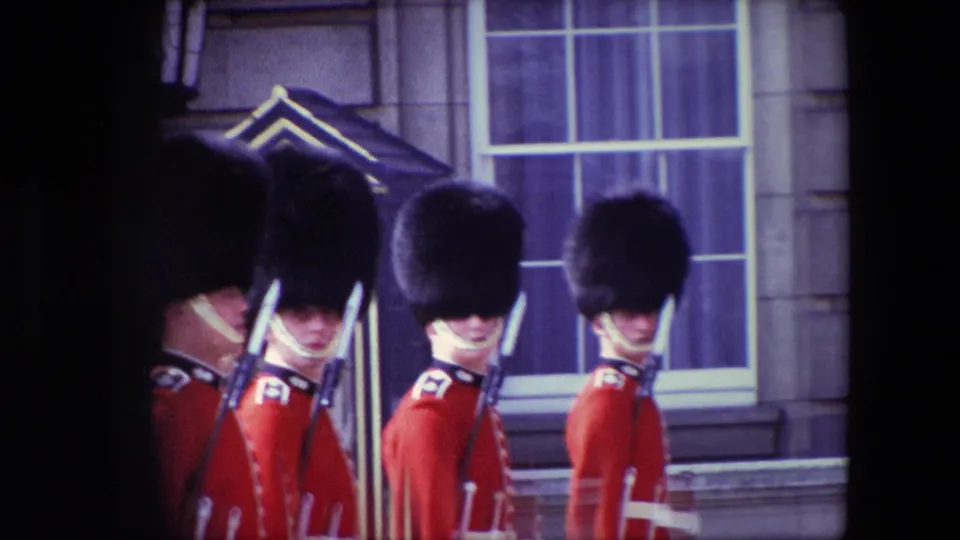 1967 London home-movie frame showing Buckingham Palace guards in red tunics and tall black caps standing at attention with rifles near palace windows.