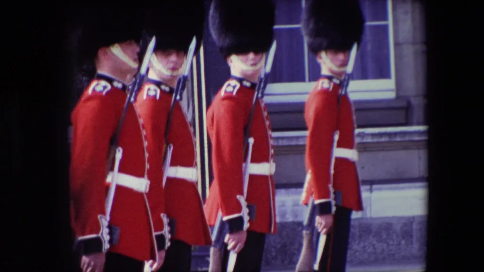 1967 London frame of several Buckingham Palace guards in ceremonial uniforms standing in profile with rifles and bayonets beside a stone exterior.