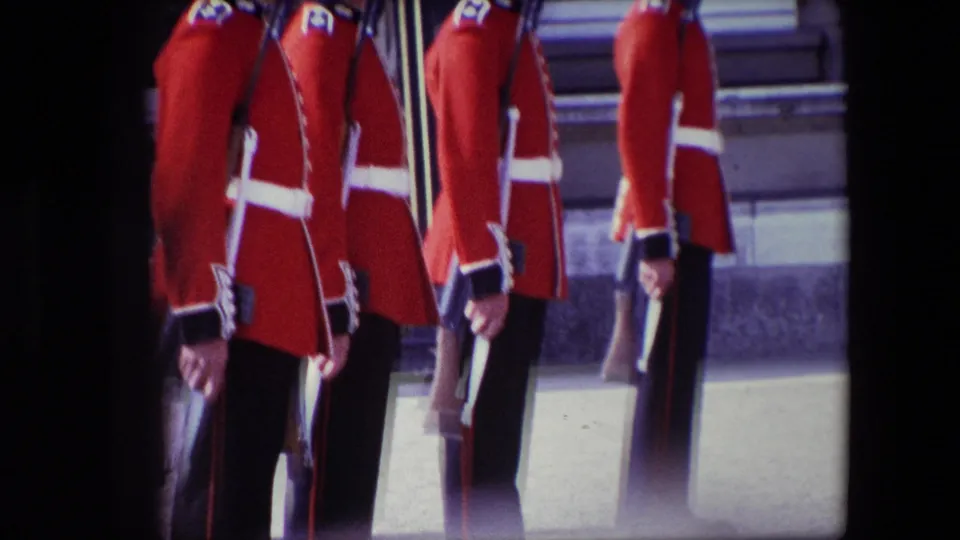 1967 London home-movie frame showing a closer, slightly shadowed view of Buckingham Palace guards in red ceremonial dress holding rifles at attention.