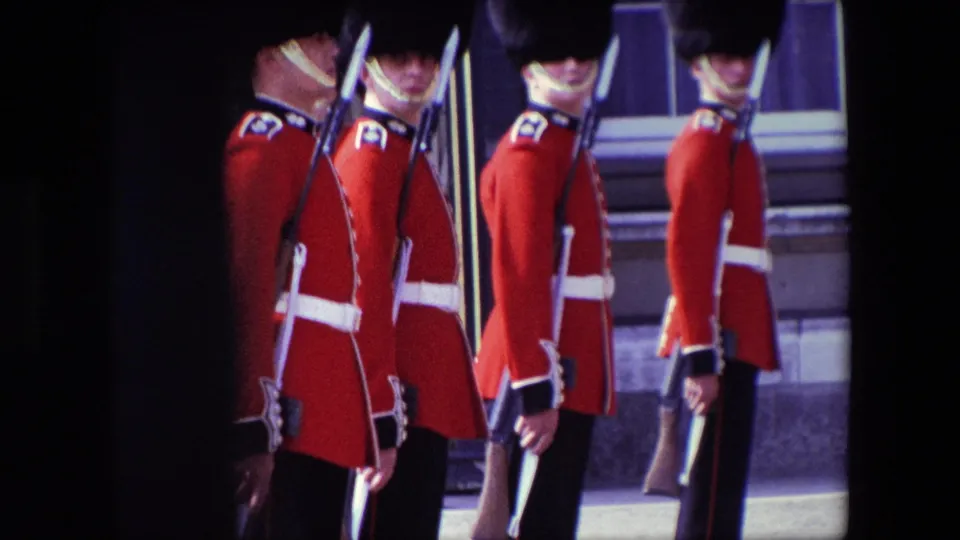 1967 London frame showing Buckingham Palace guards in red uniforms and tall black caps standing still with rifles in front of windows and stonework.
