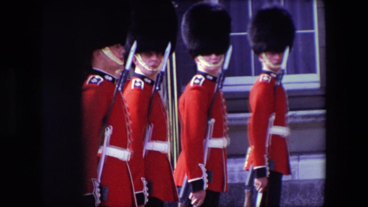 1967 London home-movie frame showing British ceremonial guards in red tunics and tall dark hats standing in formation outside a stone building.