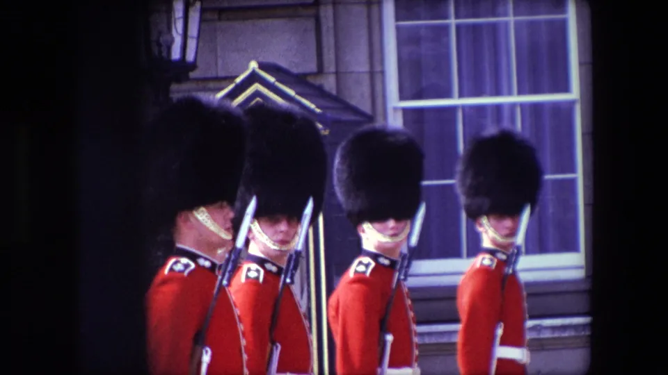 British ceremonial guards in red uniforms and bearskin-style hats appear lined up outside a London building in this 1967 8mm frame.