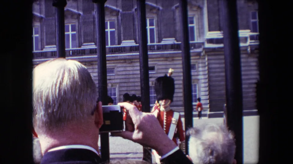 Visitor-shot 1967 London frame with tourists in the foreground photographing a ceremonial guard near railings and a large stone building.