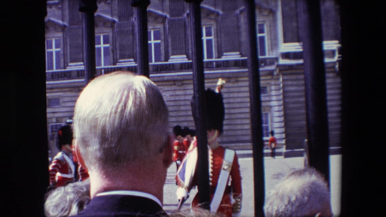In 1967 London home-movie footage, spectators stand behind black iron railings while red-coated ceremonial soldiers gather across a paved forecourt before a formal stone facade.
