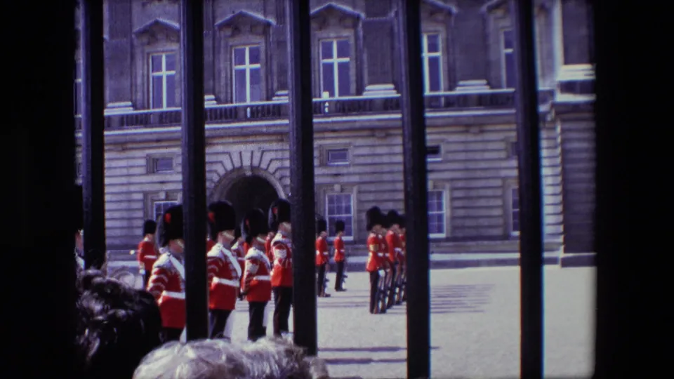 Red-coated soldiers in tall black hats stand in formation near an arched doorway, viewed through iron railings from a spectator position in London.