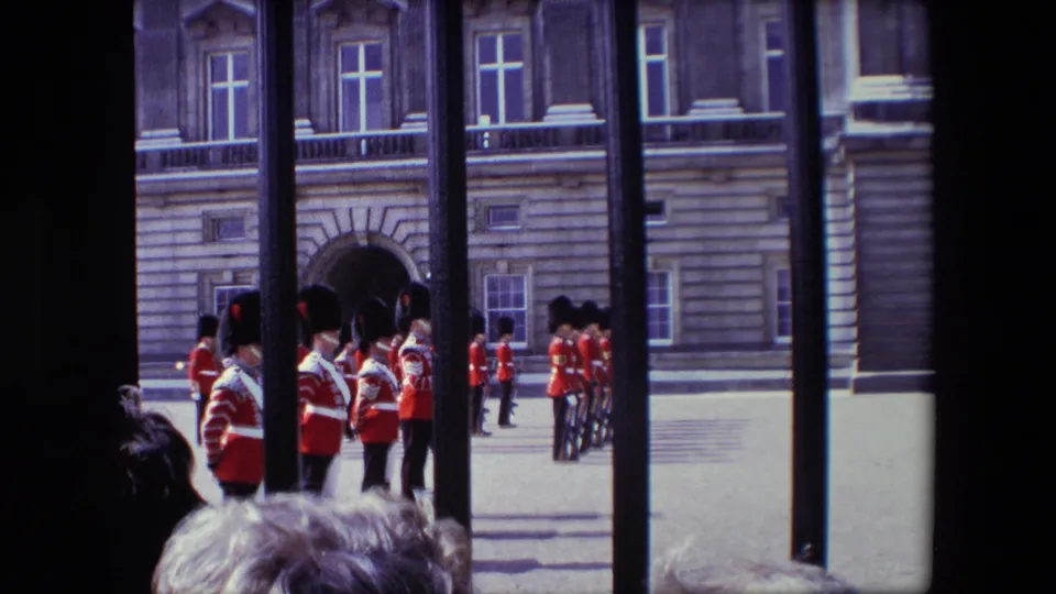 The camera frames a line of red-coated ceremonial soldiers beyond black railings, with spectators' heads in the foreground and a formal stone building behind them.