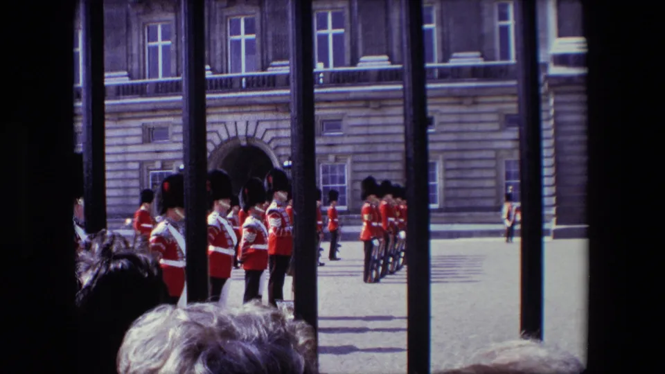 Red-uniformed ceremonial soldiers stand near an arched entrance in a London forecourt, filmed from behind railings with onlookers partly visible at the bottom of the frame.