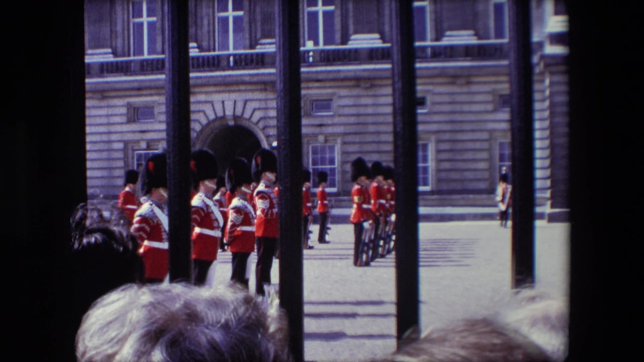 1967 London home-movie frame showing spectators behind an iron gate watching Queen's Guards in red tunics standing in a palace courtyard.