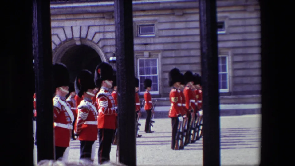 1967 London frame with Queen's Guards in red uniforms and bearskin hats lined up inside a palace courtyard, seen through a dark gate.