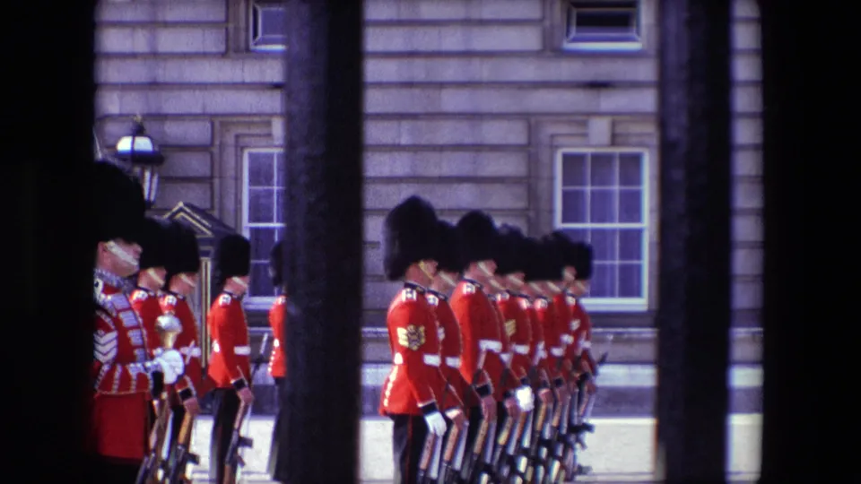 1967 London home-movie view of a line of Queen's Guards with rifles, partly obscured by iron gate bars and set against palace windows.