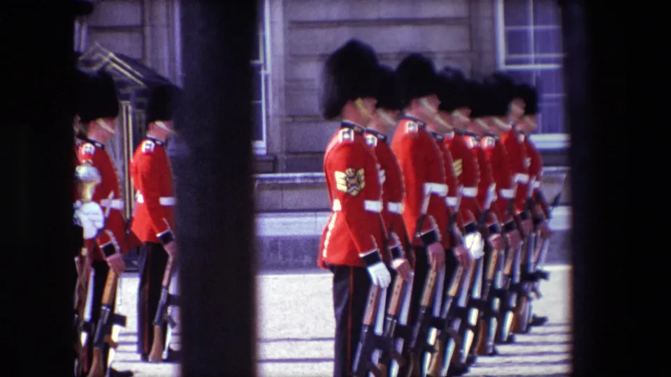 1967 London frame showing Queen's Guards in red tunics and bearskin hats standing in a close line with rifles beside a palace facade.