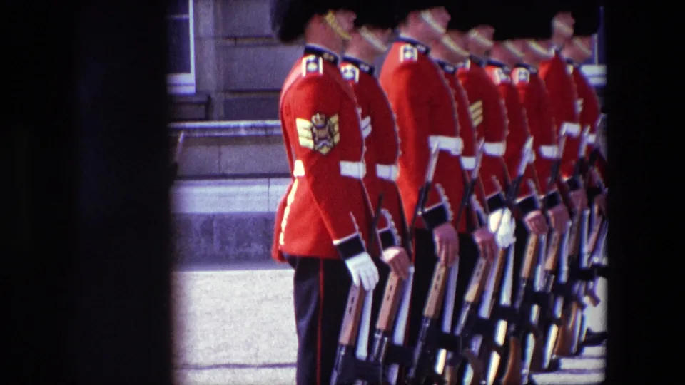 1967 London home-movie frame showing guards in red ceremonial uniforms, dark headgear, white belts and gloves, standing in formation with rifles near stone steps.