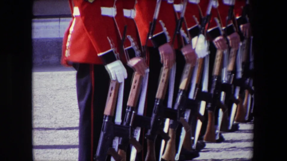 Close 1967 London frame focusing on the lower bodies of ceremonial guards in red tunics, white gloves, dark trousers, and rifles held vertically at their sides.