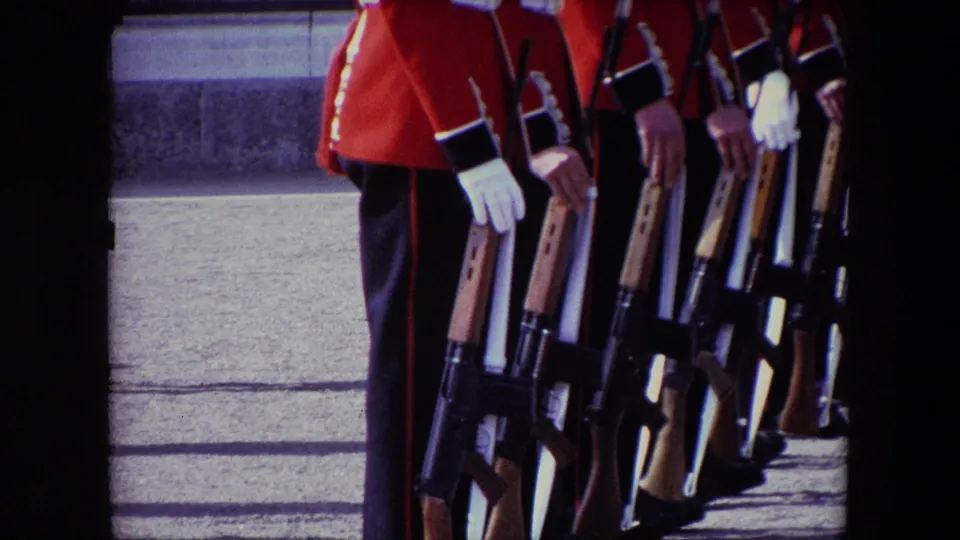 Visitor-shot 1967 London frame showing a close row of guards in red dress jackets and dark trousers, with rifles held at rest beside stone steps.