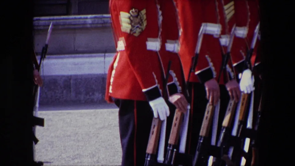 Cropped 1967 London frame with red-uniformed guards, white cross belts, gloves, and rifles, filmed close enough to show jacket sleeve insignia but not clear identities.