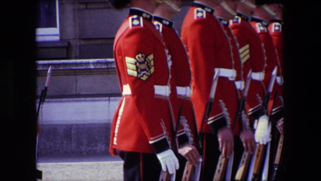 1967 London home-movie frame showing British palace guards in red ceremonial tunics and tall black hats standing in a formal line beside a stone facade.