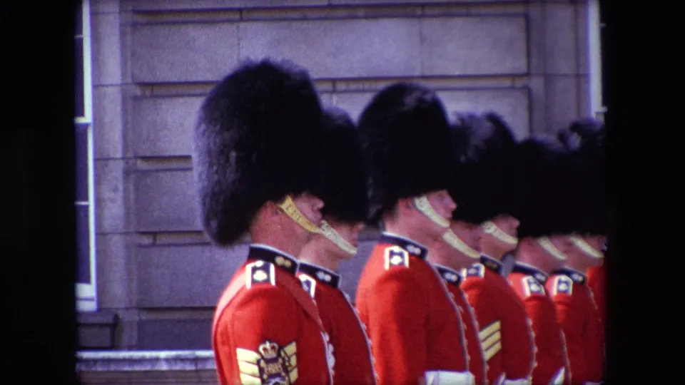 Side view of British palace guards in 1967 London, wearing red ceremonial uniforms and tall black hats while standing in close formation outside a stone building.