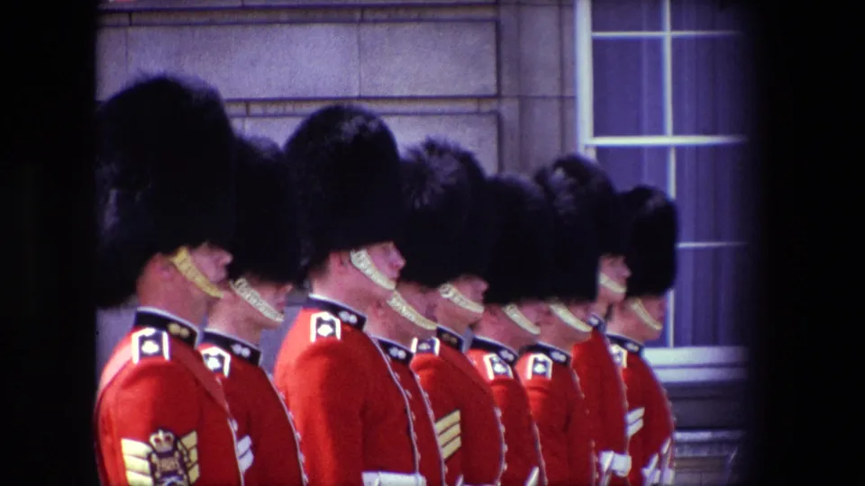 1967 London 8mm frame of a row of palace guards in red coats and black hats, partially obscured by dark film borders and viewed against a palace-like stone exterior.