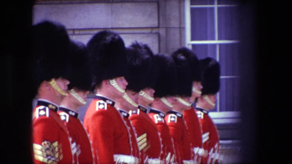 1967 London home-movie frame showing multiple British palace guards standing shoulder to shoulder in red ceremonial uniforms, with rifles and a stone building visible behind them.