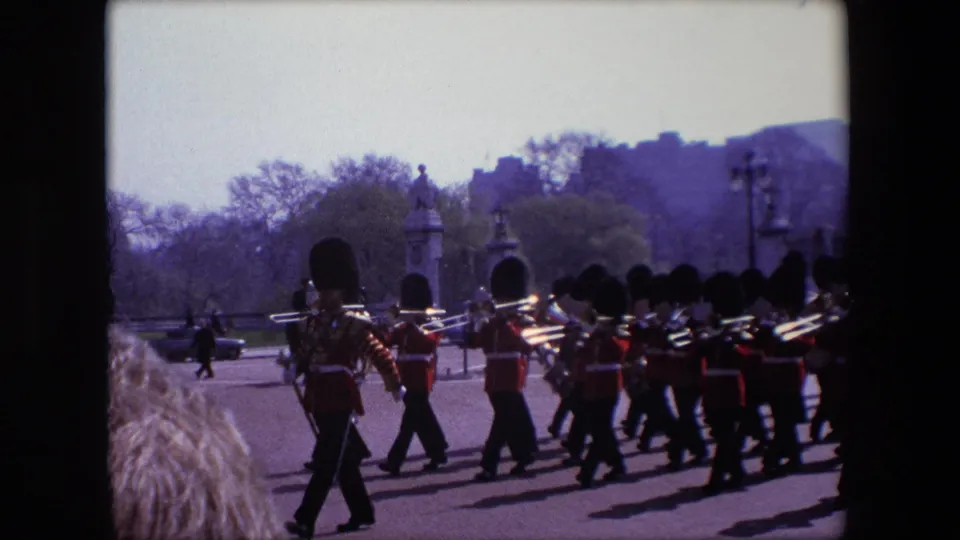 A London ceremonial band in guard uniforms passes the camera, with several trombones or brass instruments visible and park trees, lamps, and formal gates behind them.