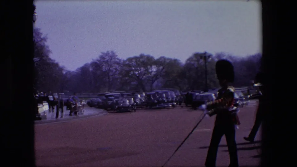 A guard in ceremonial uniform stands or marches near the foreground while rows of parked period cars, scattered pedestrians, trees, and a wide paved area fill the background.