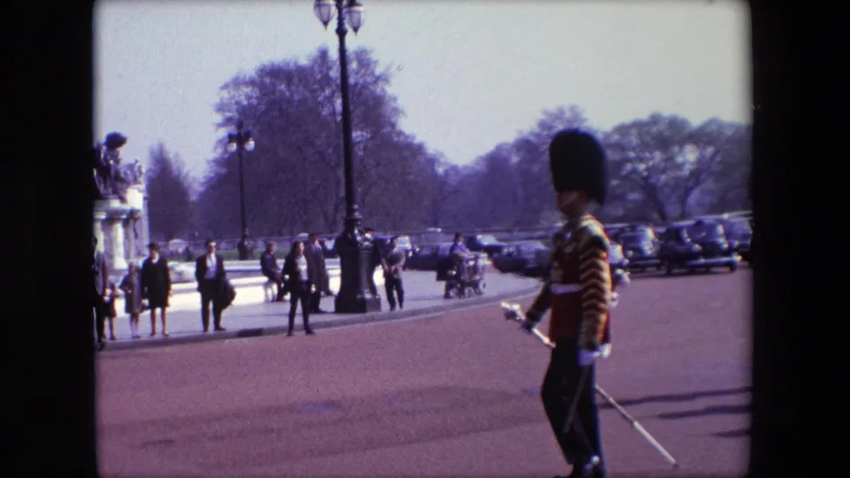 A 1967 London home-movie frame shows a ceremonial guard in red uniform at the right edge, with spectators, a tall lamppost, parked cars, trees, and formal stone sculpture or gate elements in the background.