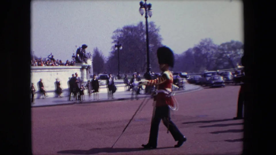 1967 London home-movie frame showing a guard in red ceremonial dress marching near the Victoria Memorial as spectators and parked cars line the background.