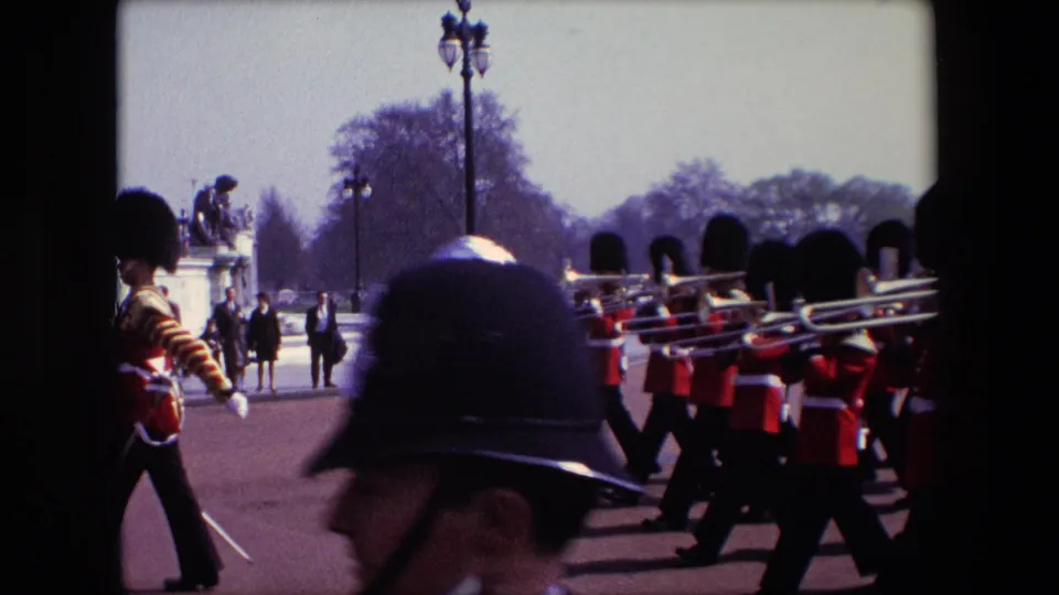 1967 London home-movie frame with Queen's Guard musicians carrying brass instruments near Buckingham Palace, partly blocked by a helmeted person in the foreground.