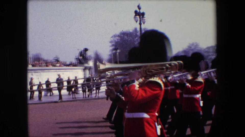 1967 London frame showing ceremonial band members with trombones marching close to the camera, with spectators and the Victoria Memorial visible behind them.