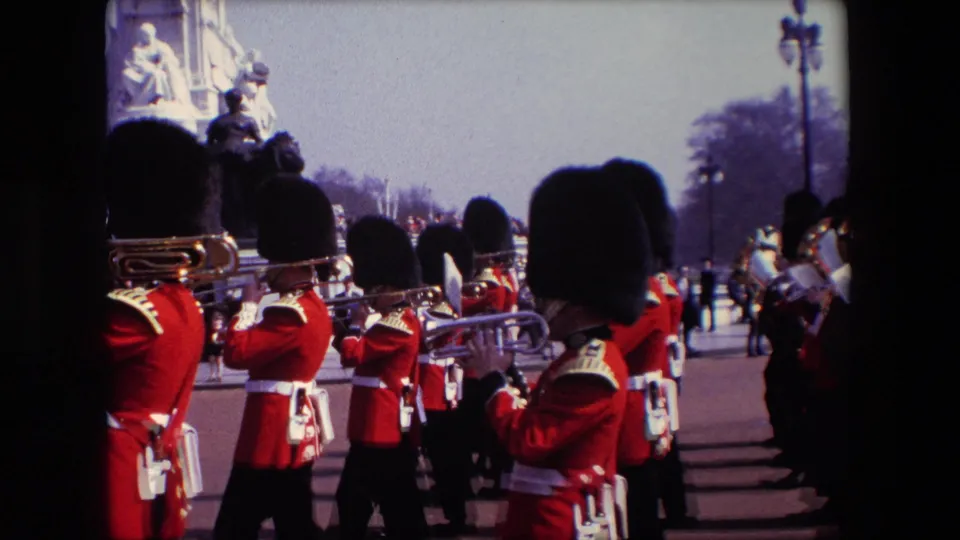 1967 London home-movie frame of a ceremonial guard band marching near Buckingham Palace, with red uniforms, brass instruments, spectators, and the memorial setting in view.