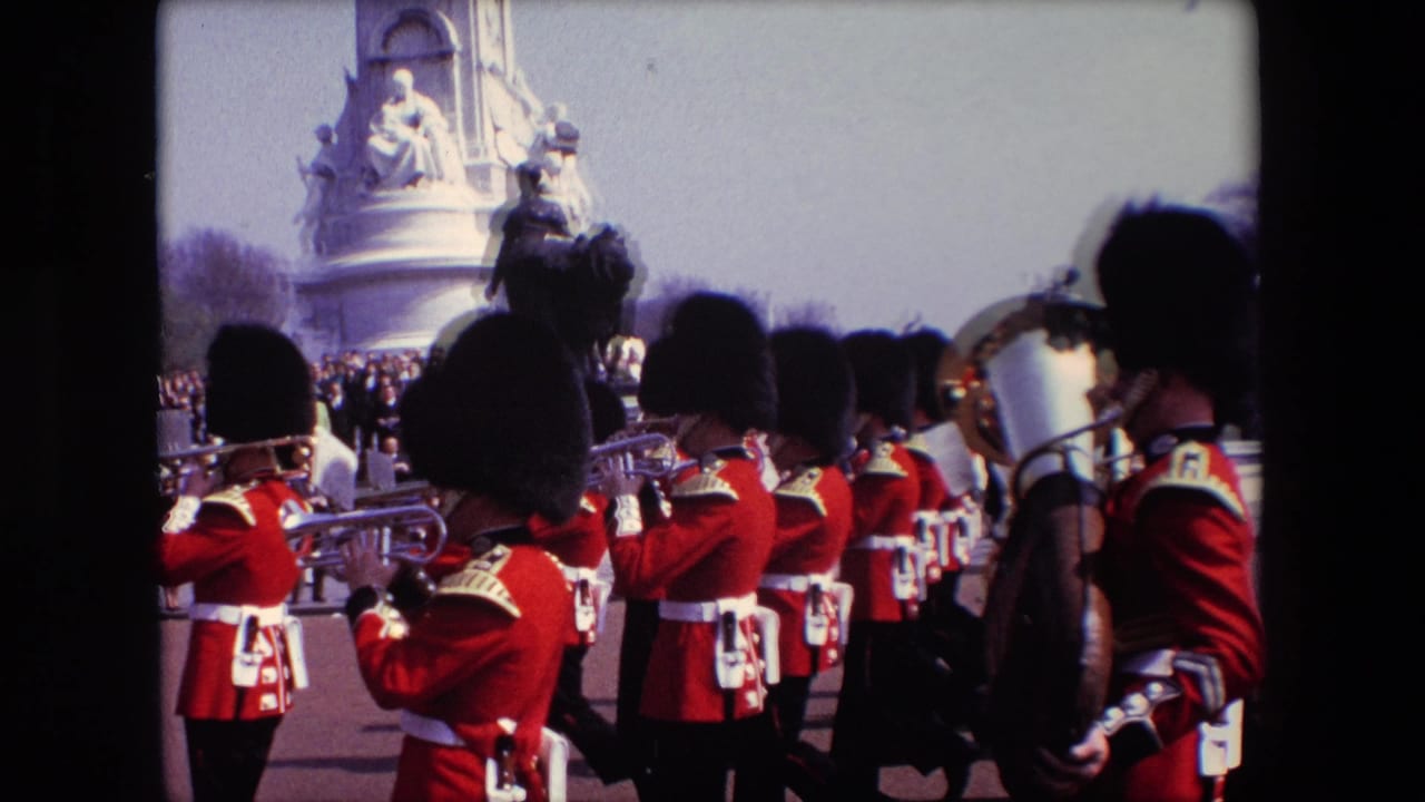 Uniformed band members in red coats and tall black hats march near a large stone monument.