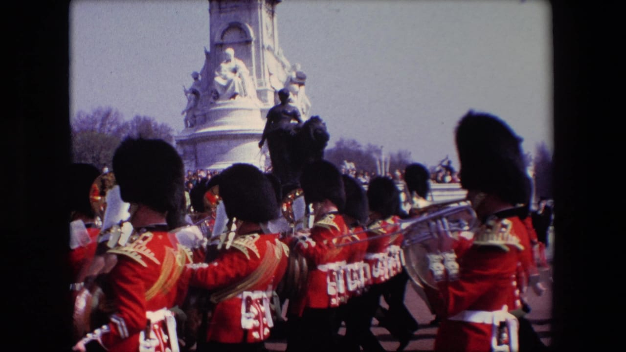 Marching musicians in red uniforms play brass instruments with a monument behind them.