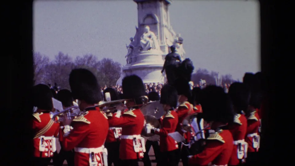 1967 London home-movie frame with red-uniformed band members and brass instruments crossing the foreground as spectators stand around a large stone monument.