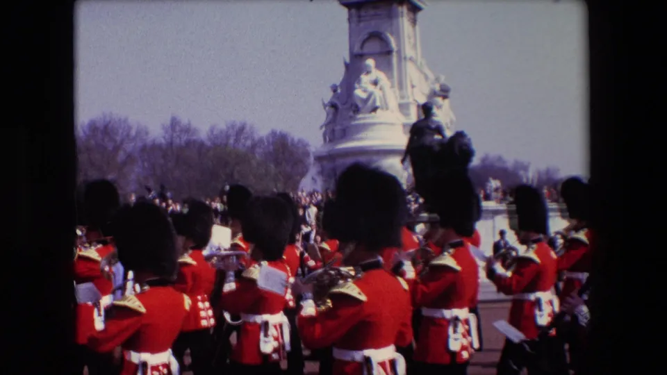 1967 London frame showing ceremonial musicians in red uniforms and tall hats clustered near spectators, with a large stone monument and trees behind them.