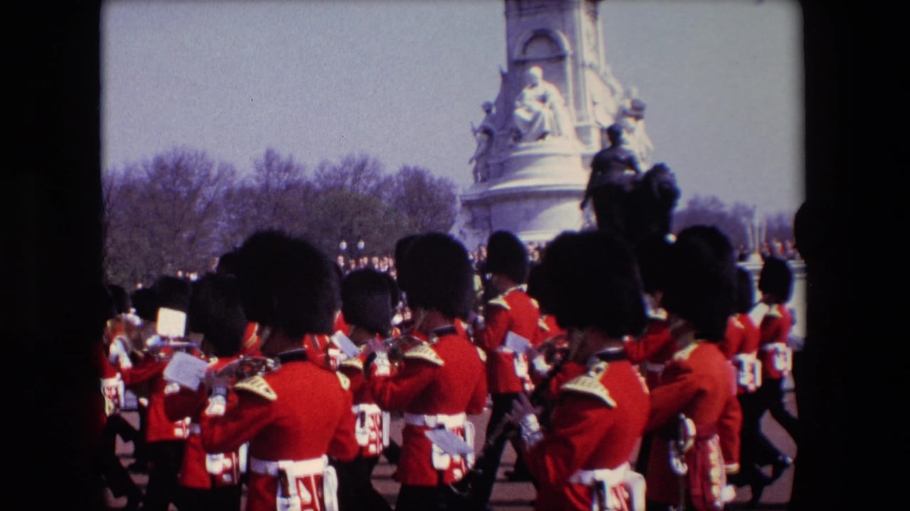 1967 London home-movie frame showing Queen's Guard soldiers in red tunics and tall black hats marching in formation near a large stone monument and spectators.