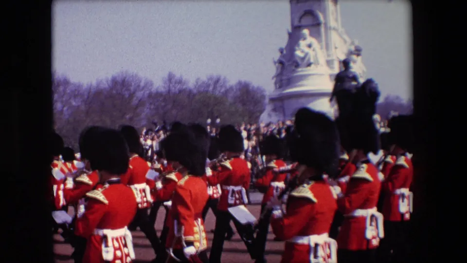 1967 London frame of Queen's Guard soldiers moving across the parade route in red ceremonial uniforms, with a crowd, bare trees, and part of a stone memorial visible behind them.