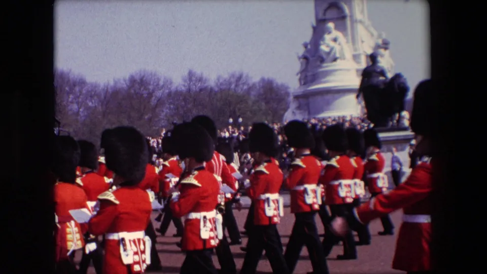 1967 London home-movie view from close to the procession, with several Queen's Guard soldiers in red coats and tall black hats marching past spectators near the monument area.