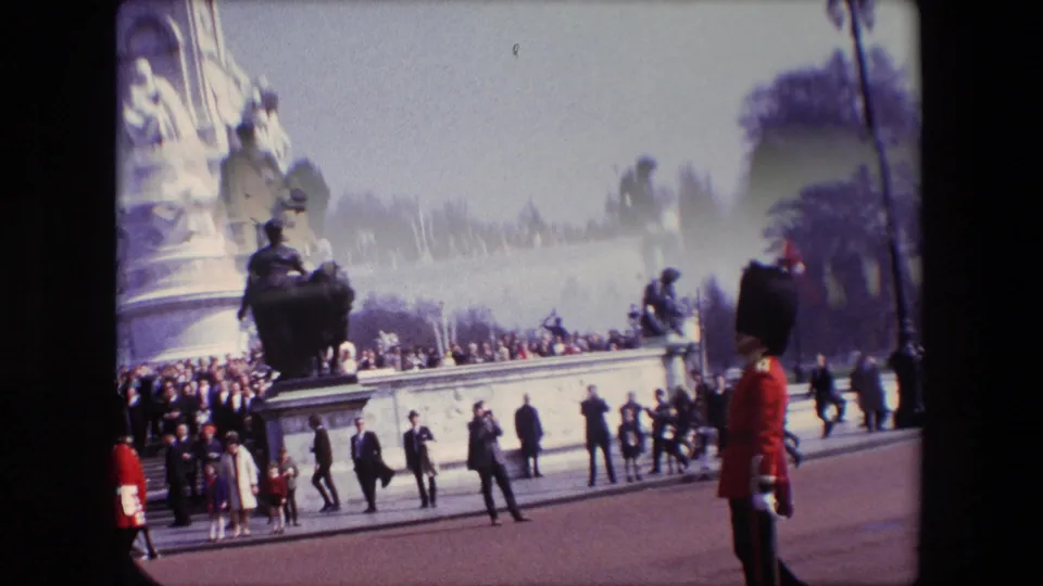 1967 London home-movie frame with a wider tilted view of a ceremonial area, showing a guard in red at the right, spectators along the stone monument, and people walking across the paved space.